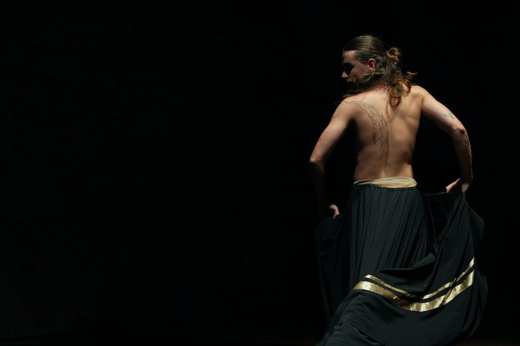 Tono Ferriol dancing on a dark stage, seen from behind, in a long black skirt with gold bands